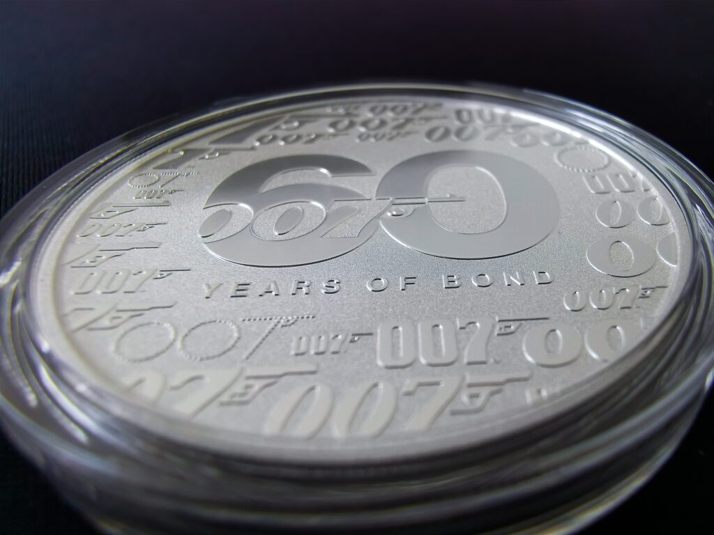 Moneda de plata por los 60 años del agente 007 con el número 60 al centro y 007 sobrepuesto. Alrededor de la moneda se observa 007 en diferentes tipografías.